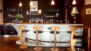 A bar area with a counter, stools, hanging lights, shelves with bottles, and framed pictures on the wall. - Hôtel La Chaloupe (Port-des-Barques)