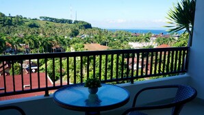 Un balcón con una mesa redonda de vidrio y dos sillas de mimbre, con vista a una zona costera con palmeras y casas.
