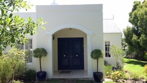 A white building with a black door, surrounded by greenery and a brick pathway.