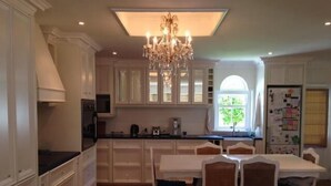 A kitchen with white cabinets, a chandelier, a dining table with chairs, and a window with a view of greenery.