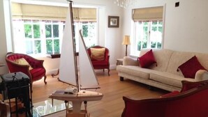 A living room with a sailboat model, red chairs, a beige sofa, and large windows with curtains.