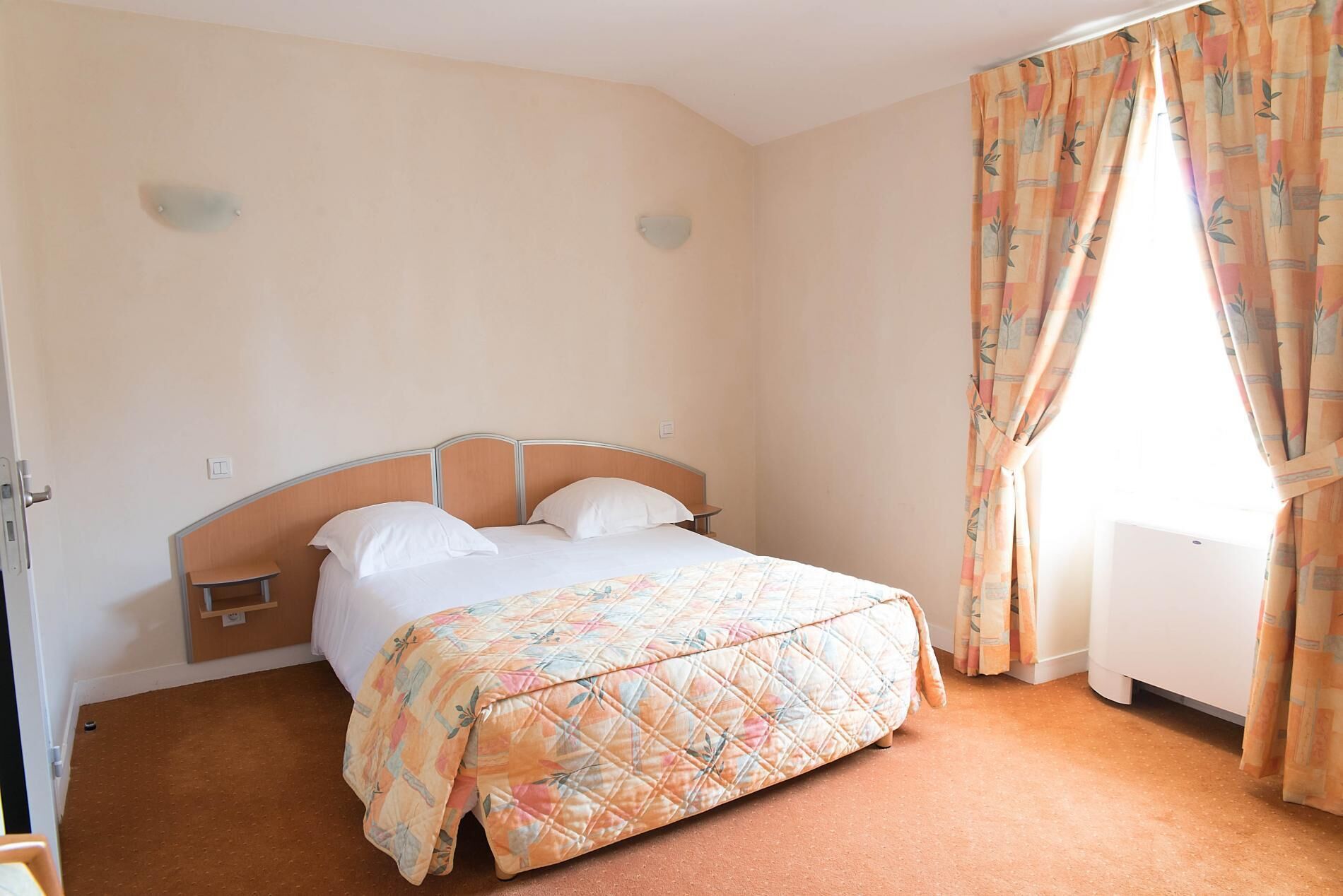 Habitación de hotel con cama, mesita de noche, lámpara de pared y ventana con cortinas.