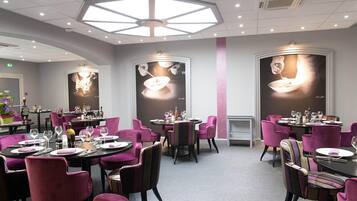 Un restaurante con mesas redondas y sillas moradas, que presenta una obra de arte de gran tamaño en la pared y un tragaluz.