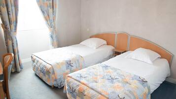 A room with two single beds, each with a floral-patterned bedspread. There is a small round table between the beds, and a chair is visible to the left.