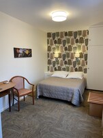 A hotel room with a bed, a chair, a desk, and a wall with a tree pattern.