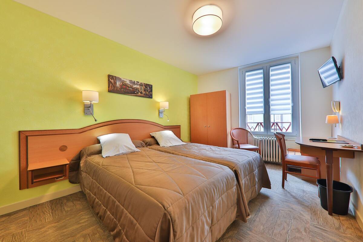 A hotel room with two beds, a desk, a chair, a TV, and a window with blinds.
