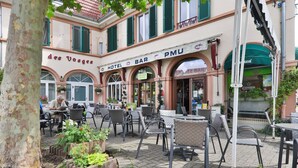 Ein Außenbereich eines Cafés mit Tischen, Stühlen, einem Baum und einem Gebäude mit der Aufschrift "des Vosges HOTEL BAR PMU".