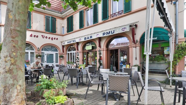 An outdoor cafe area with tables and chairs, a tree, and a building with a sign that reads "des Vosges HOTEL BAR PMU". - HOTEL Bar des VOSGES (Munster)