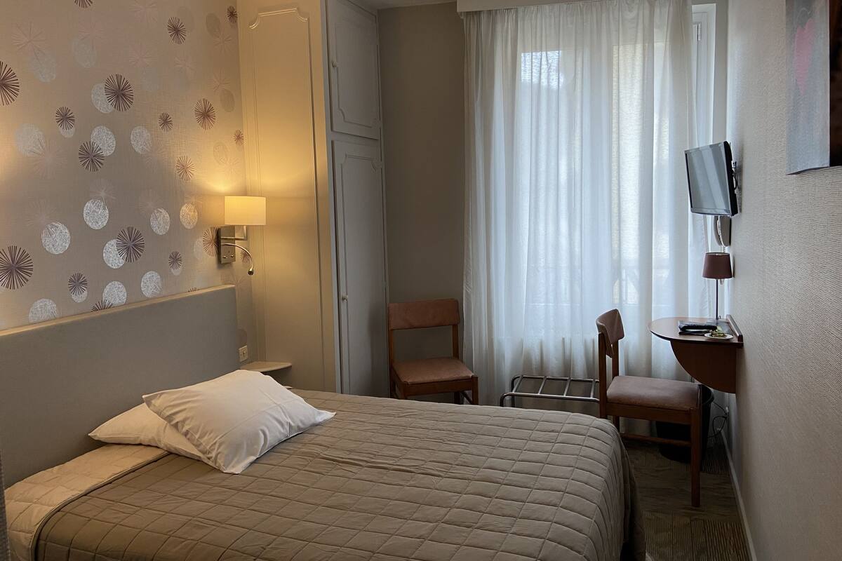 A hotel room with a bed, a desk with a chair, a TV, and a window with curtains.