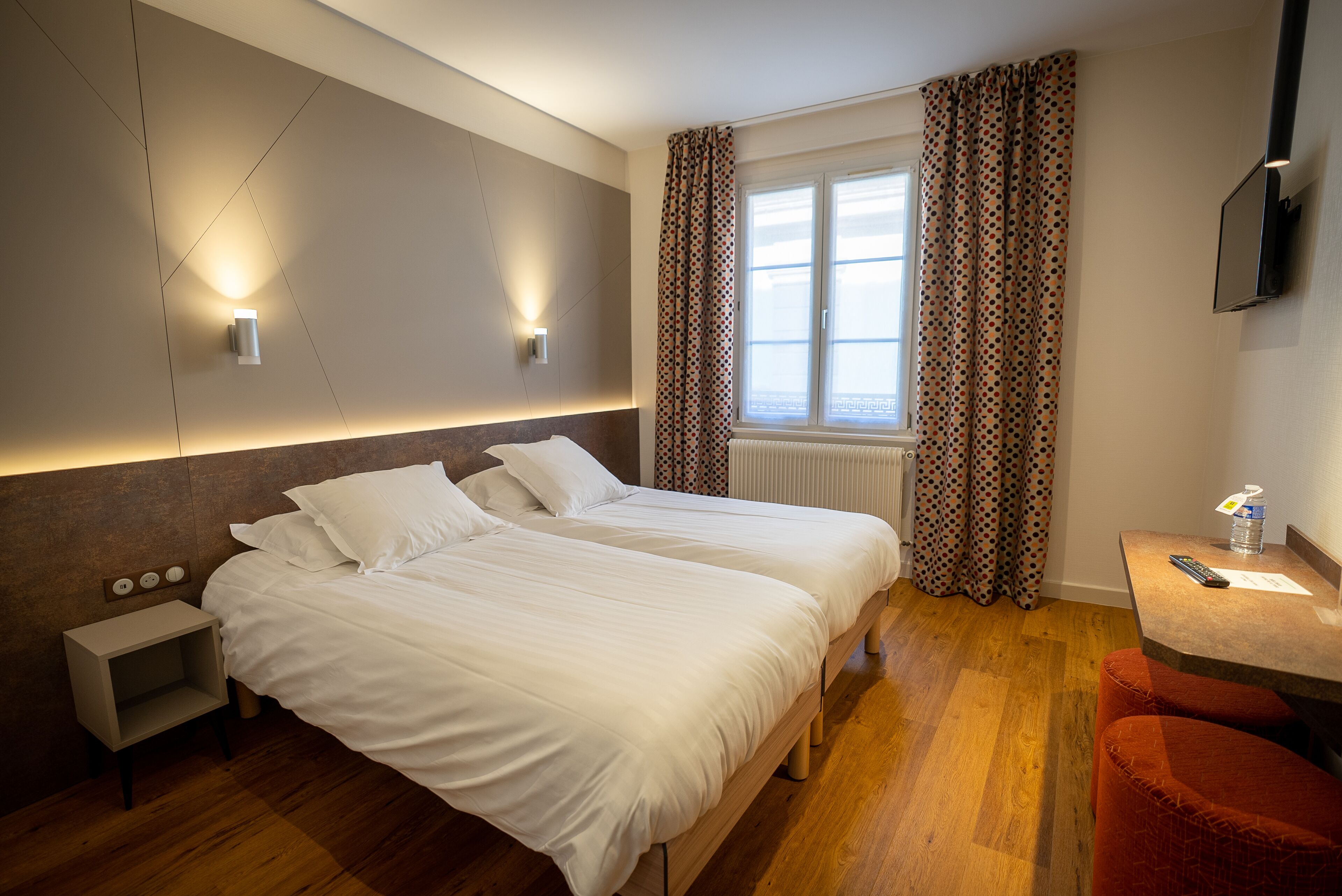 A hotel room with two beds, a small bedside table, a nightstand, a desk with a chair, a window with curtains, and a TV mounted on the wall.