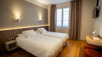 A hotel room with two beds, a small bedside table, a nightstand, a desk with a chair, a window with curtains, and a TV mounted on the wall.