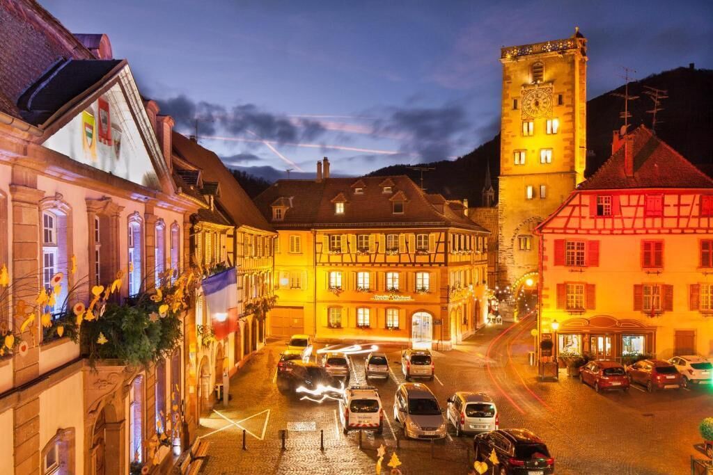 A historic street with traditional architecture, a prominent clock tower, and a row of parked cars.