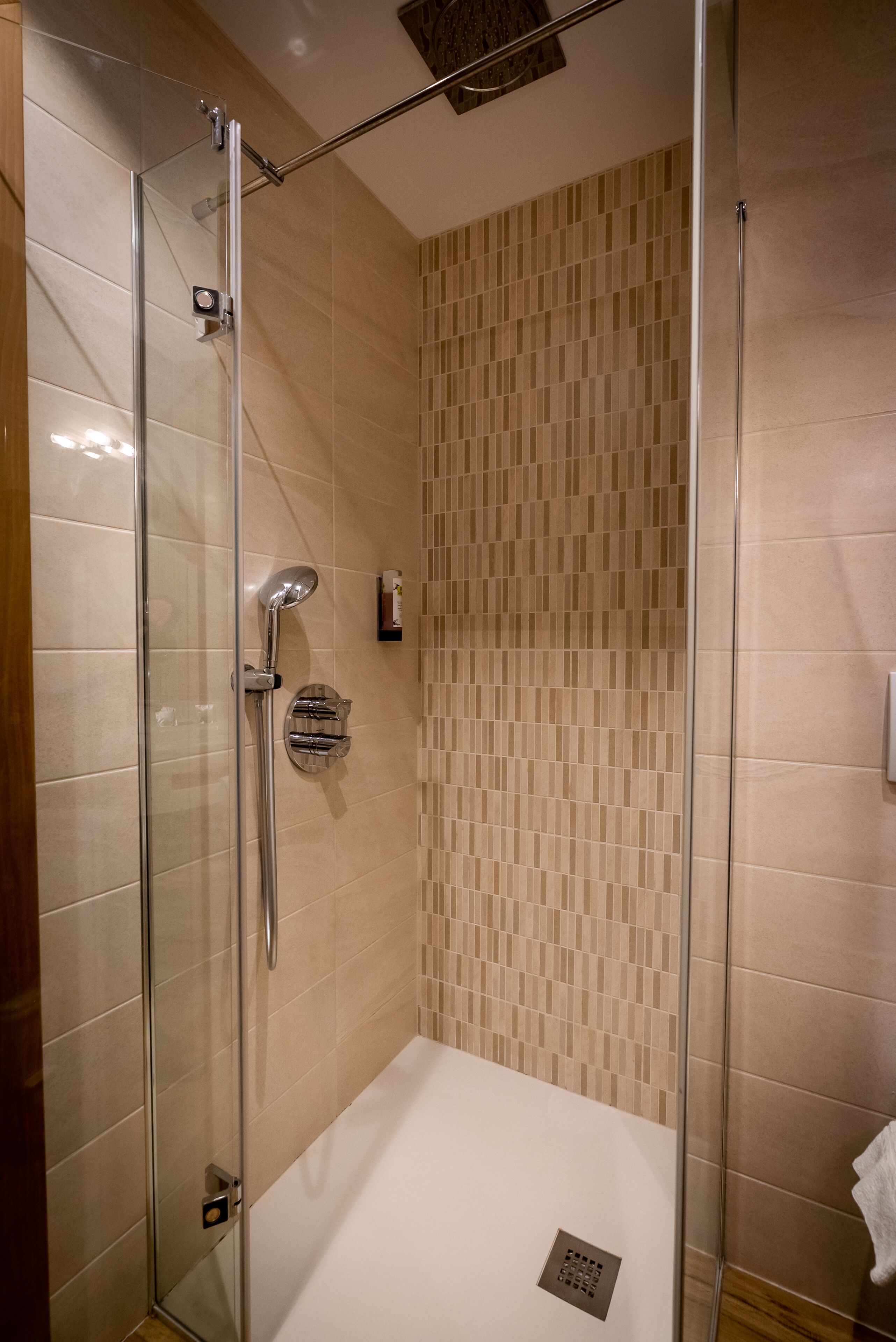 A glass-enclosed shower with a control panel, beige tiled walls, and a white bathtub.