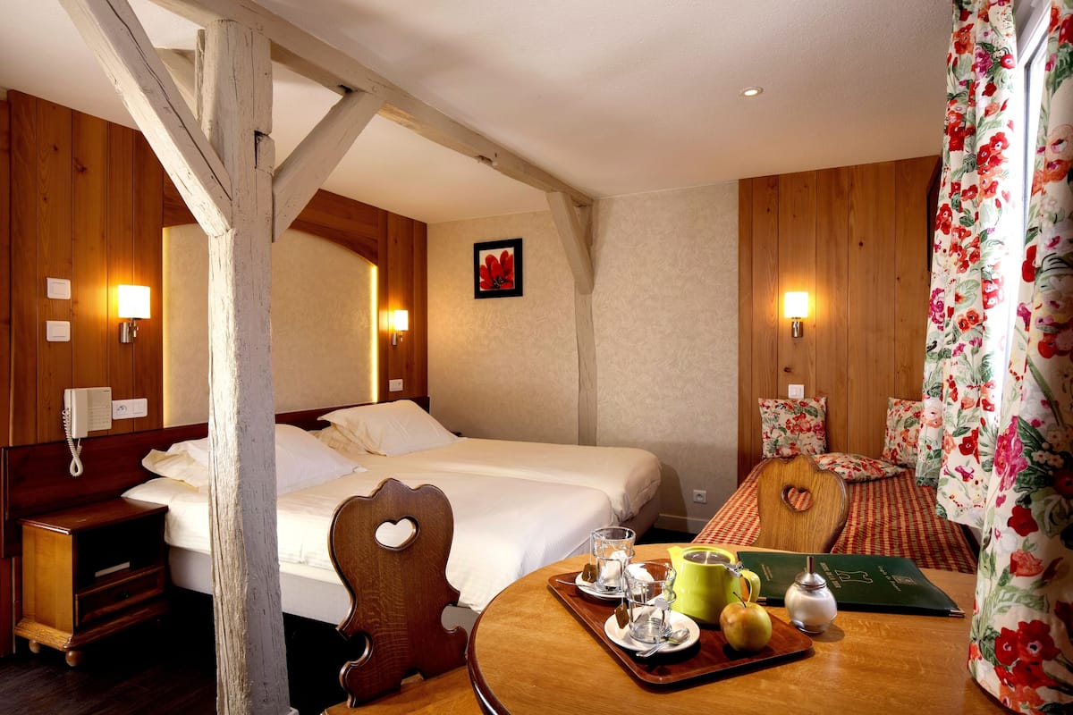 Une chambre d’hôtel avec deux lits, des murs en bois, une chaise en bois, une table avec des articles pour le petit-déjeuner et un rideau à motifs floraux.