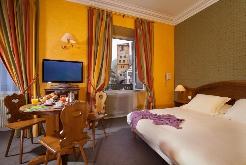 A hotel room with a bed, a desk with a television, a window with curtains, and a view of buildings outside.