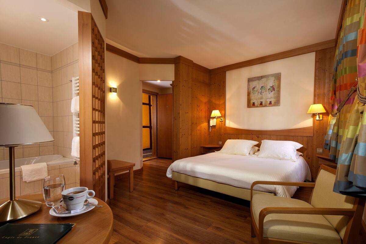 Une chambre d’hôtel avec un lit, des lampes de chevet, une chaise, une petite table avec une tasse et un verre, et un tableau accroché au mur.