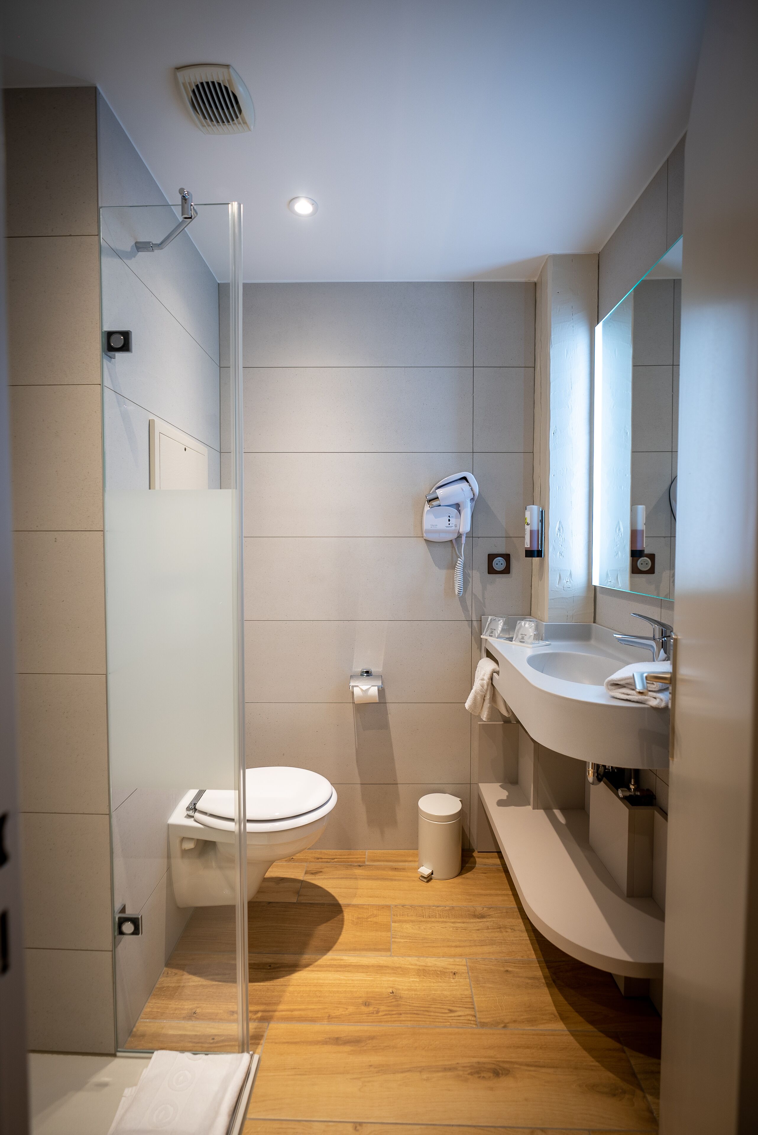 Un baño moderno con mampara de vidrio, inodoro blanco, lavabo con espejo y un cubo de basura pequeño.