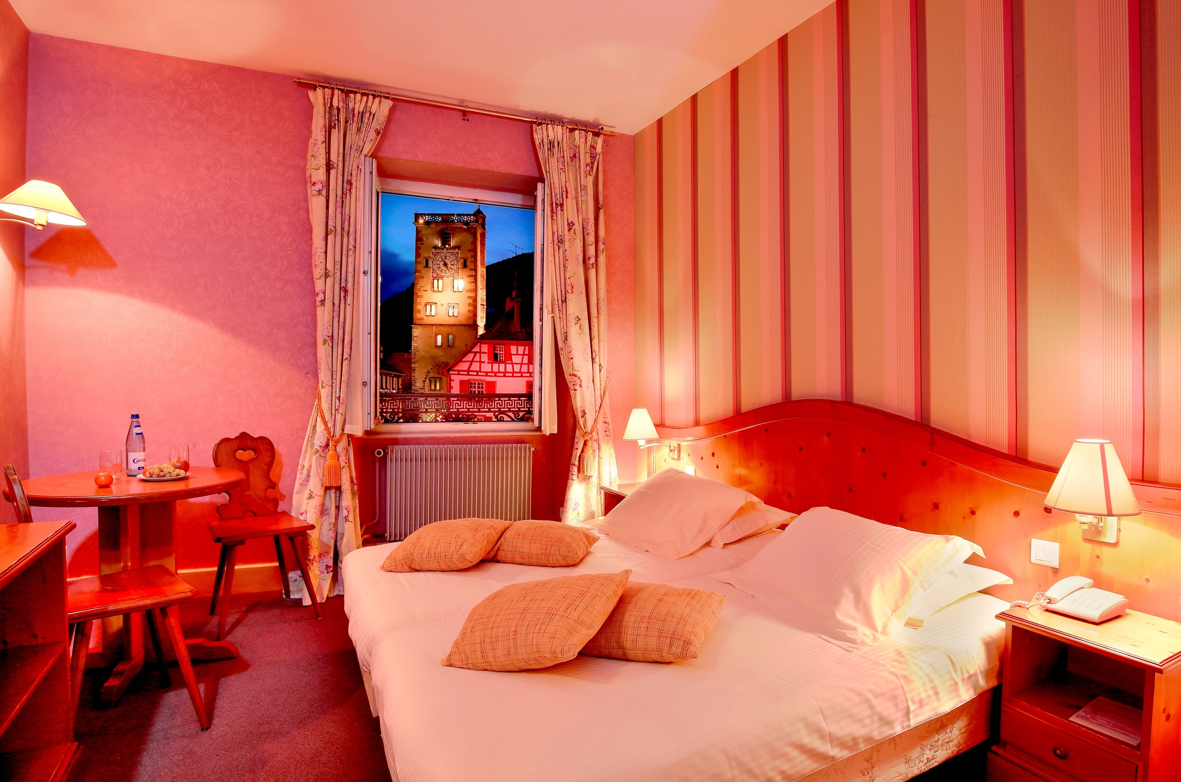 Une chambre d’hôtel avec un lit, une table de chevet, une lampe et une vue sur un immeuble par la fenêtre.