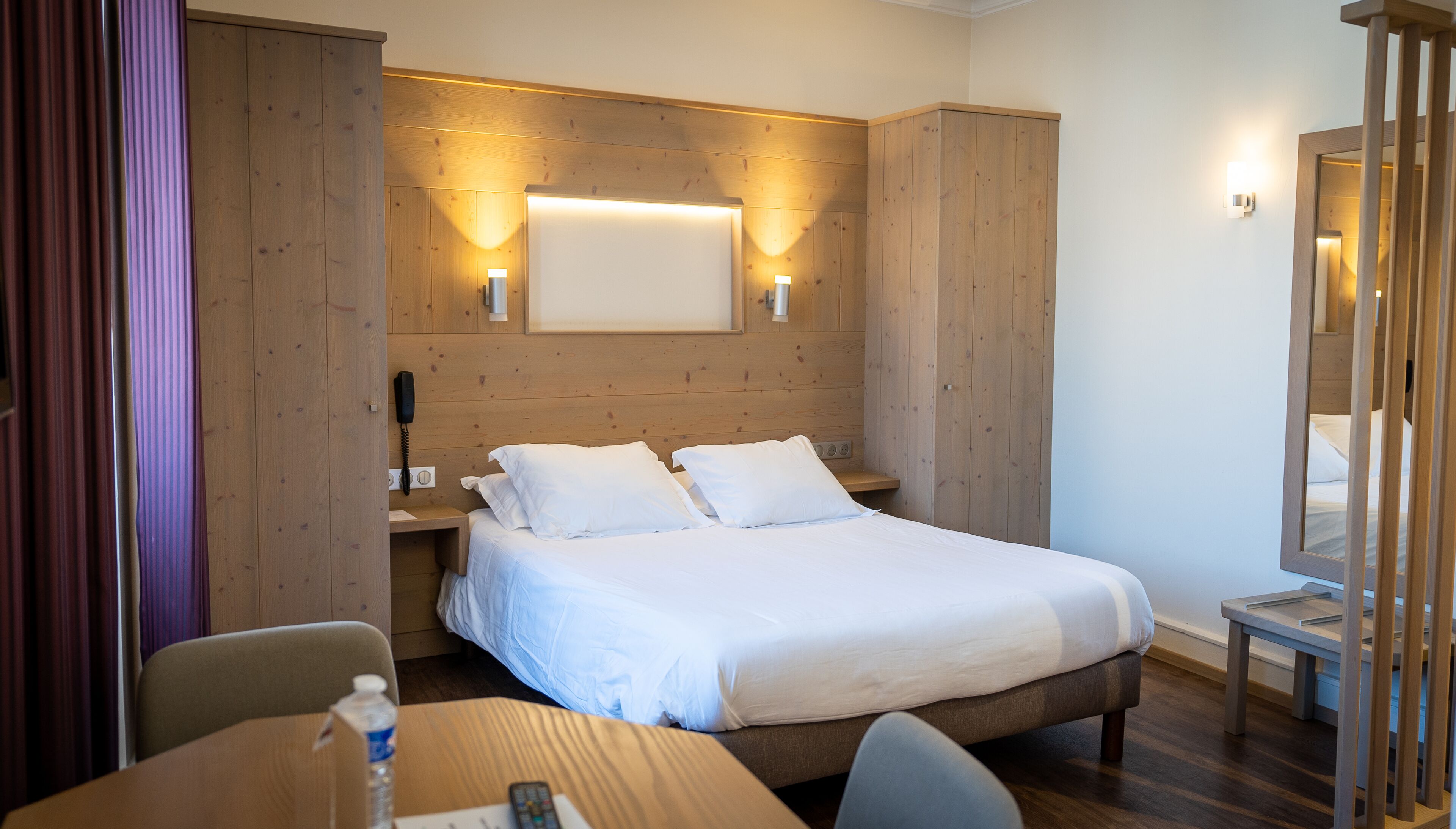 Habitació d'hotel amb capçal de llit de fusta, un llit amb roba de llit blanca, una tauleta de nit amb una làmpada, un mirall i un escriptori amb una cadira.