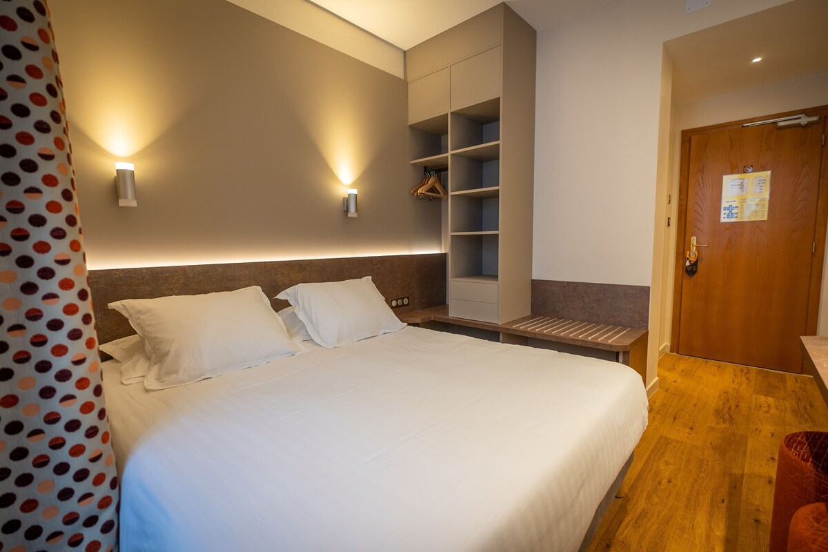 Une chambre d’hôtel comprenant un lit, deux lampes fixées au mur, un banc en bois, une armoire et une porte sur laquelle est apposée une affiche.