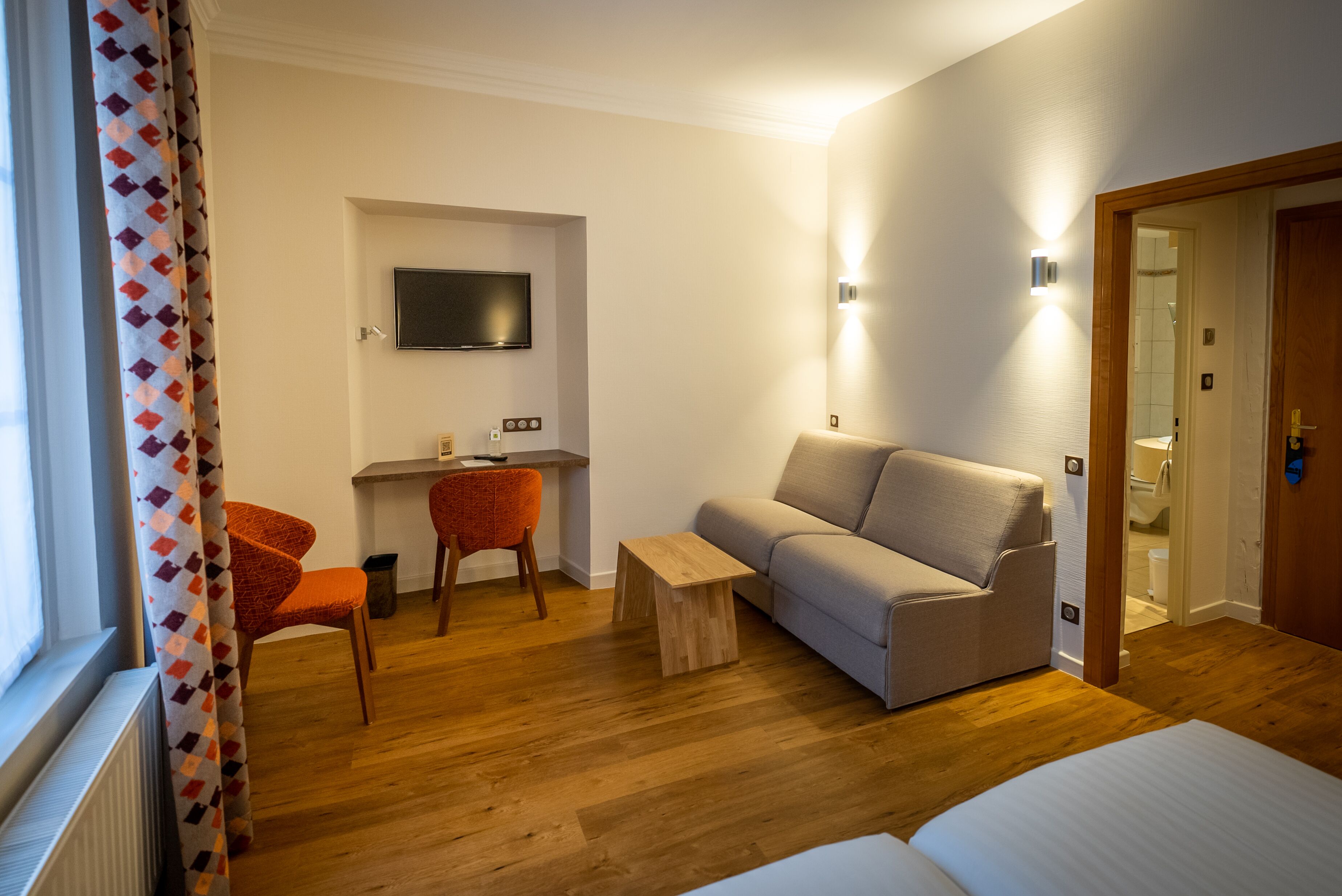 A hotel room with a wooden floor, a sofa, a small table, a TV mounted on the wall, and a door leading to a bathroom.