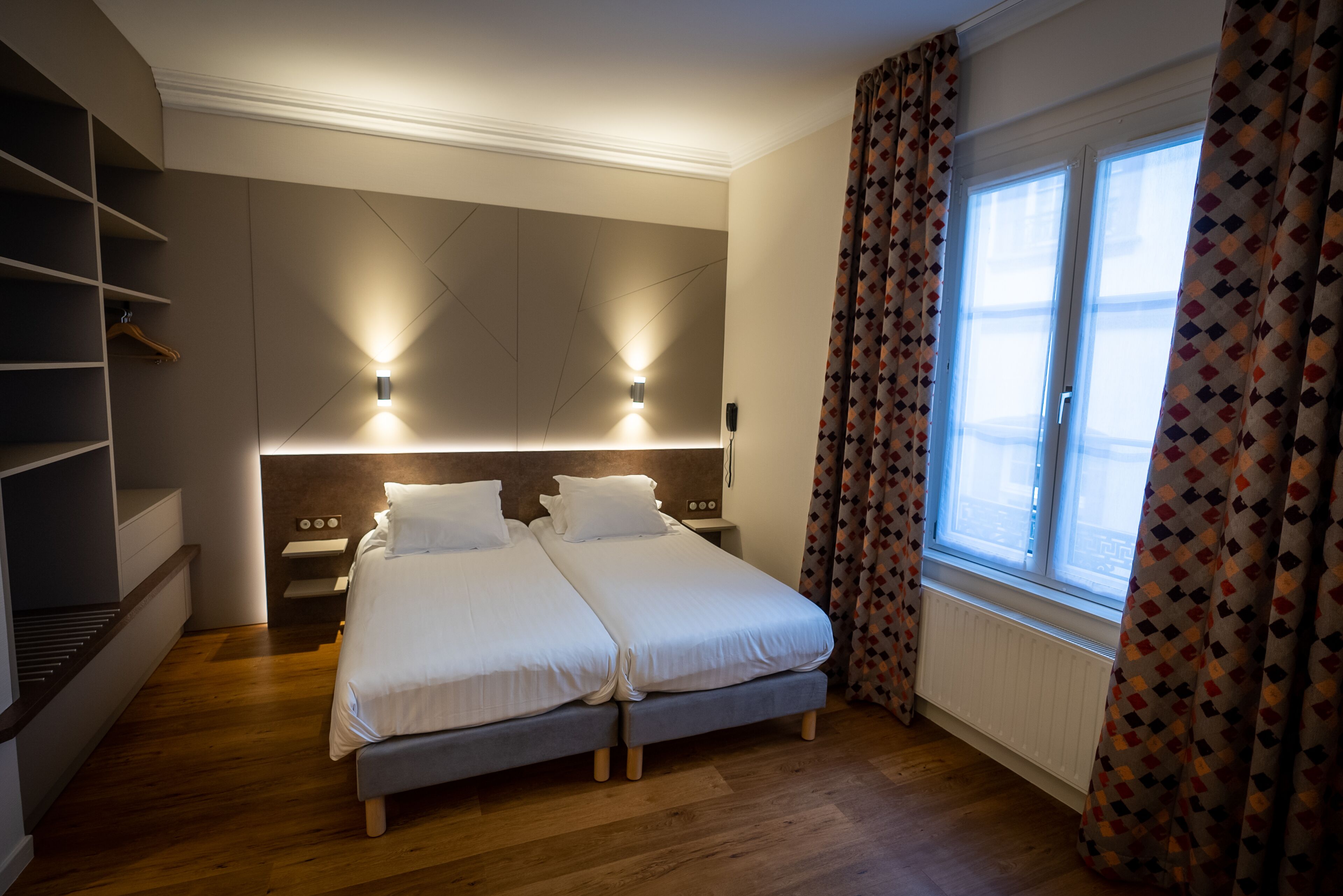 Une chambre moderne avec deux lits, des étagères intégrées, des lampes de chevet et une fenêtre agrémentée de rideaux à motifs.