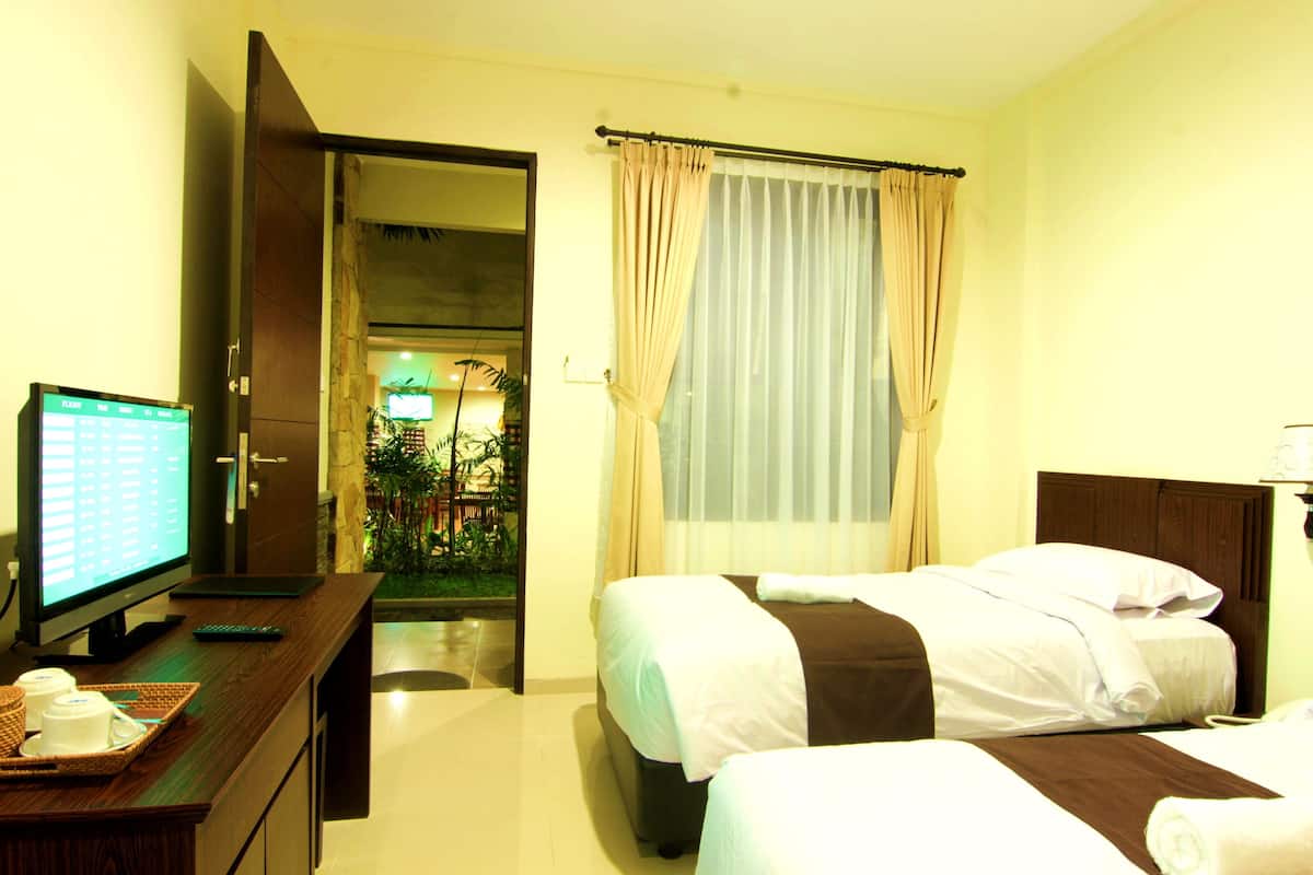A hotel room with two beds, a desk with a computer and a tray of cups, and a view of a garden through a window.