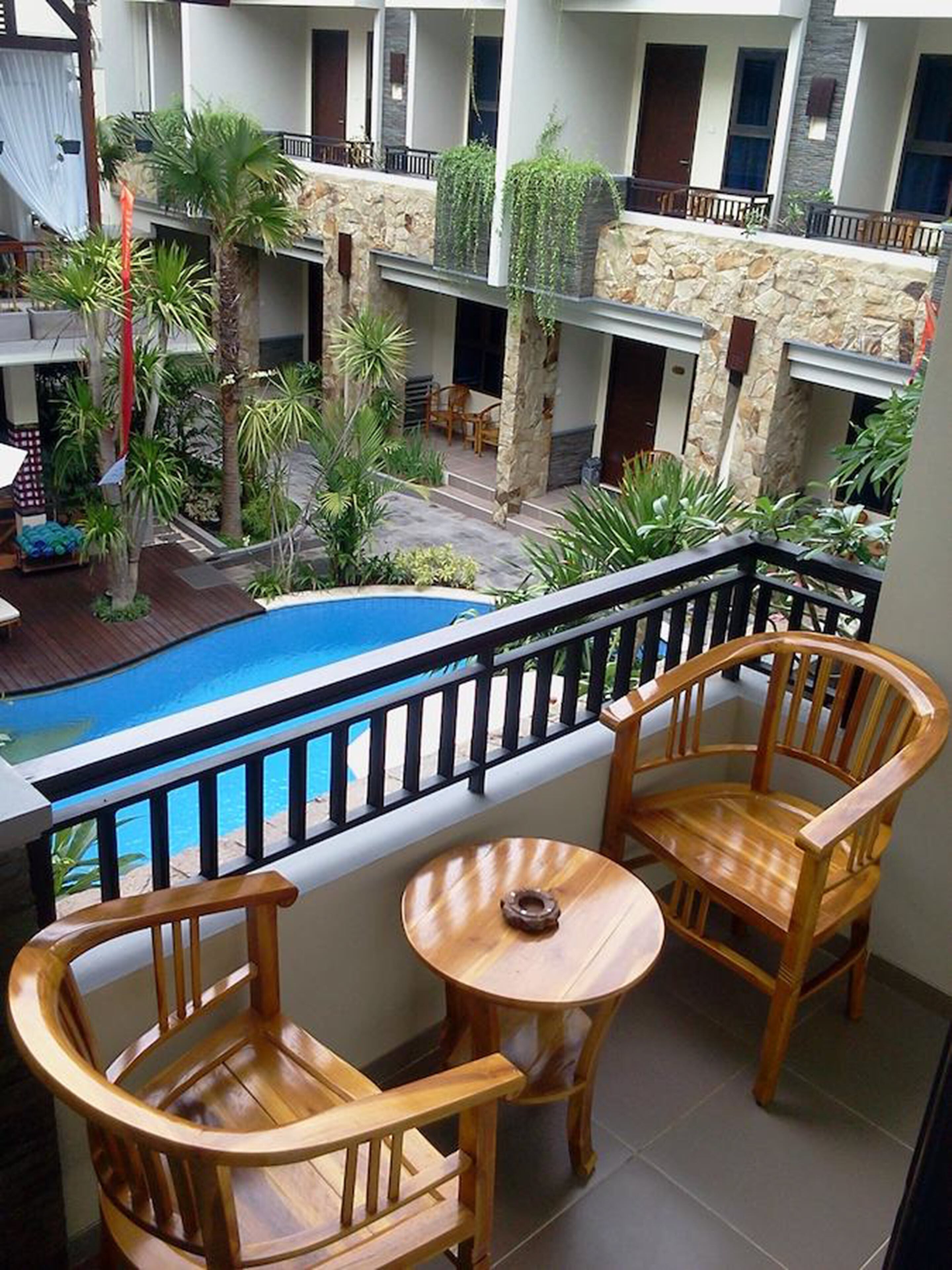 A balcony with wooden chairs and a small table, overlooking a swimming pool and a courtyard with plants and a stone wall.
