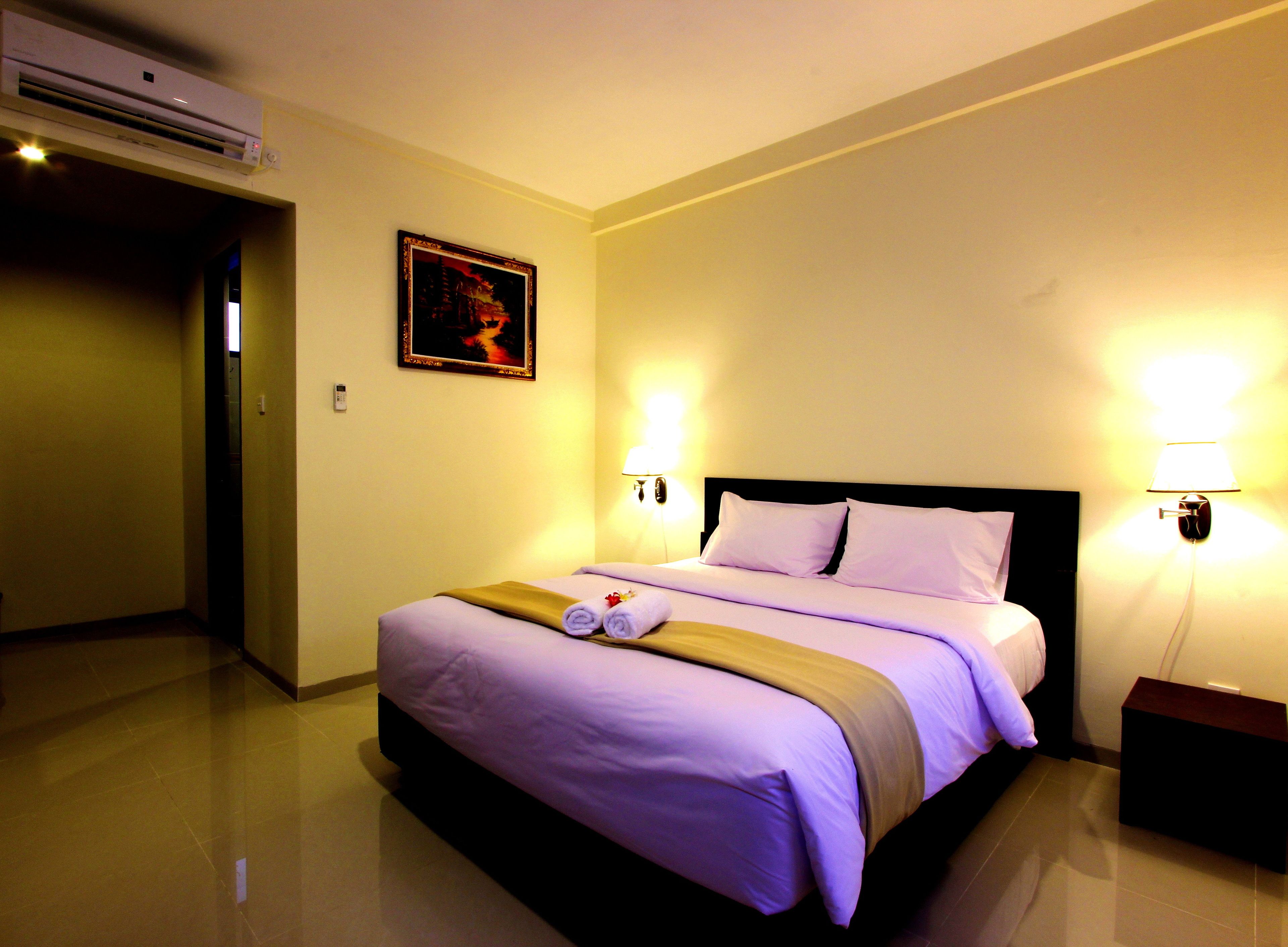 A hotel room with a bed, a wall-mounted air conditioner, a painting, and two bedside lamps.