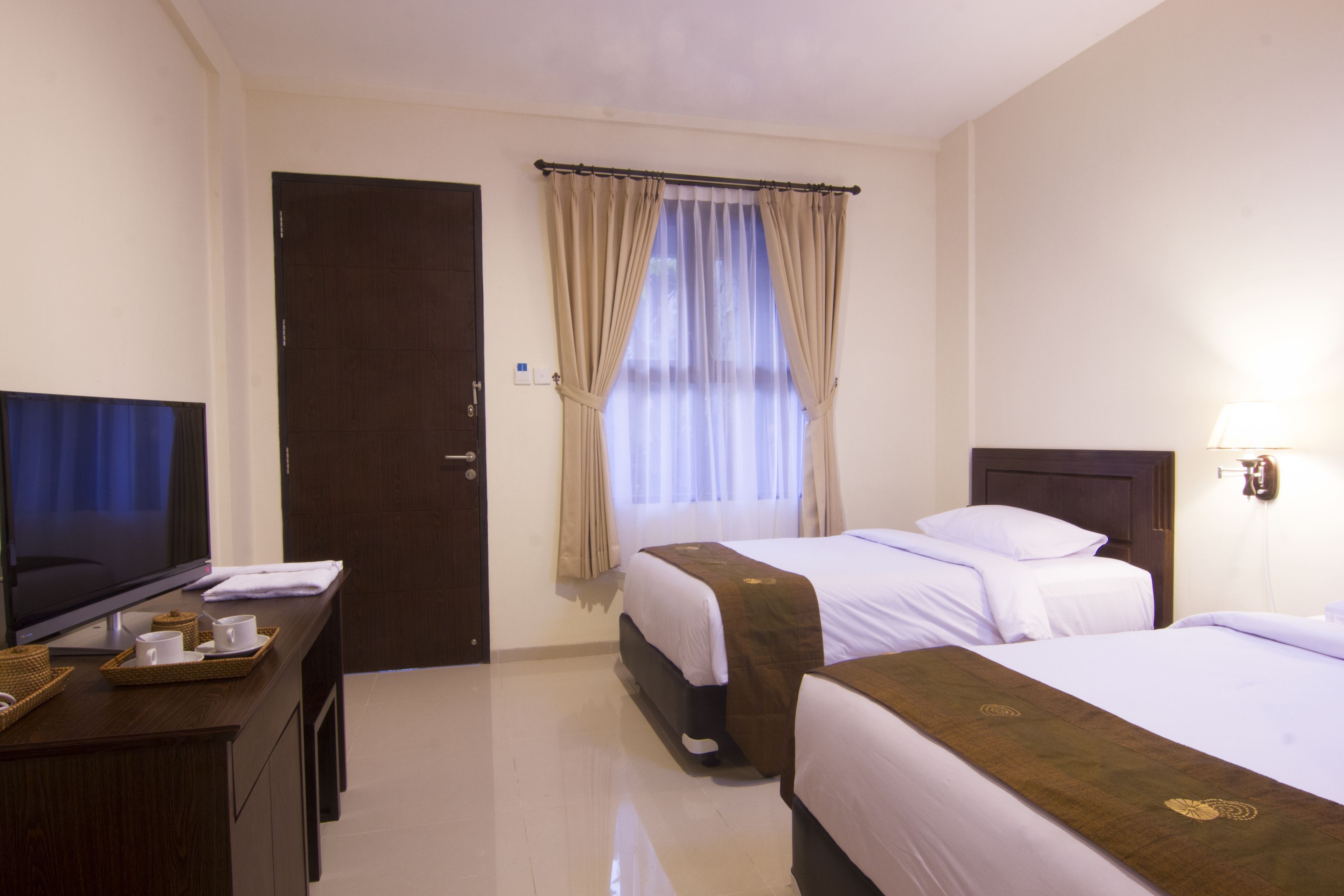 A hotel room with two beds, a TV on a desk, a window with curtains, and a lamp on the wall.