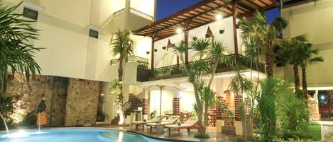 A hotel pool area with lounge chairs, palm trees, and a covered seating area.