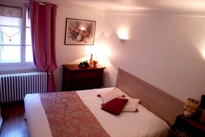 A neatly made bed with a patterned bedspread and pillows, a bedside table with a bottle and a lamp, a radiator, and a window with curtains. - Auberge du Brand (Turckheim)