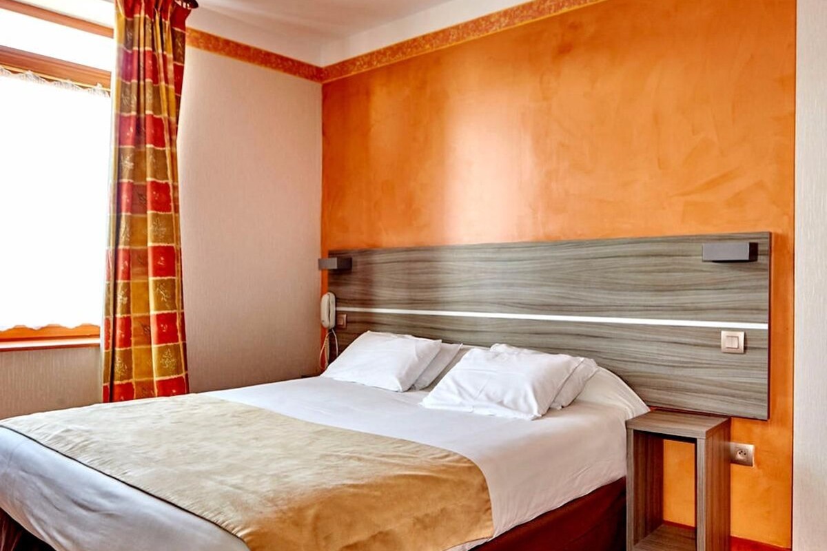 A hotel room with a large bed, a wooden headboard, a bedside table, and a window with curtains.