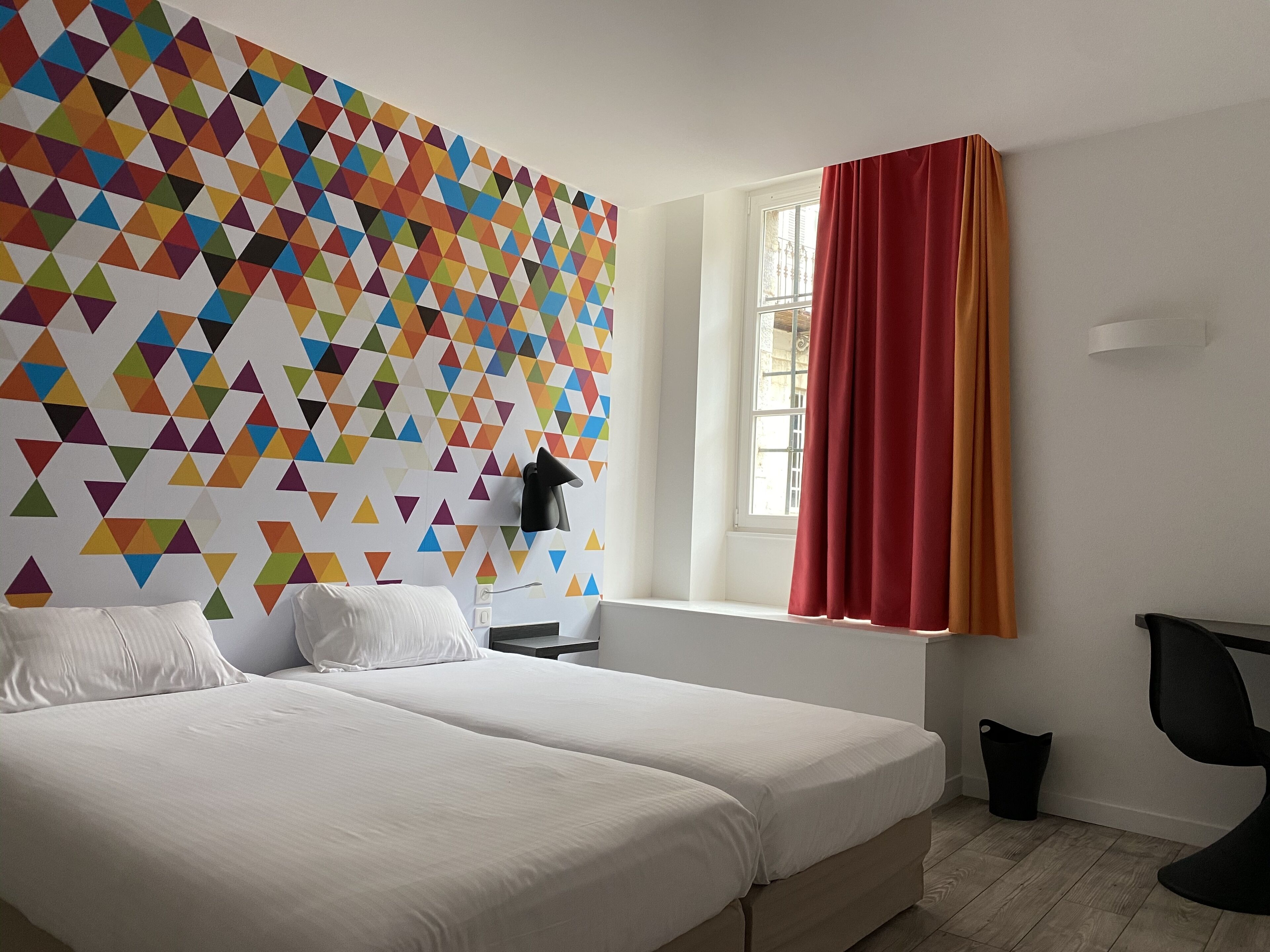 Ein Hotelzimmer mit einer großen Wand mit geometrischem Muster, einem Bett mit weißer Bettwäsche, einem Fenster mit roten Vorhängen und einem schwarzen Stuhl.