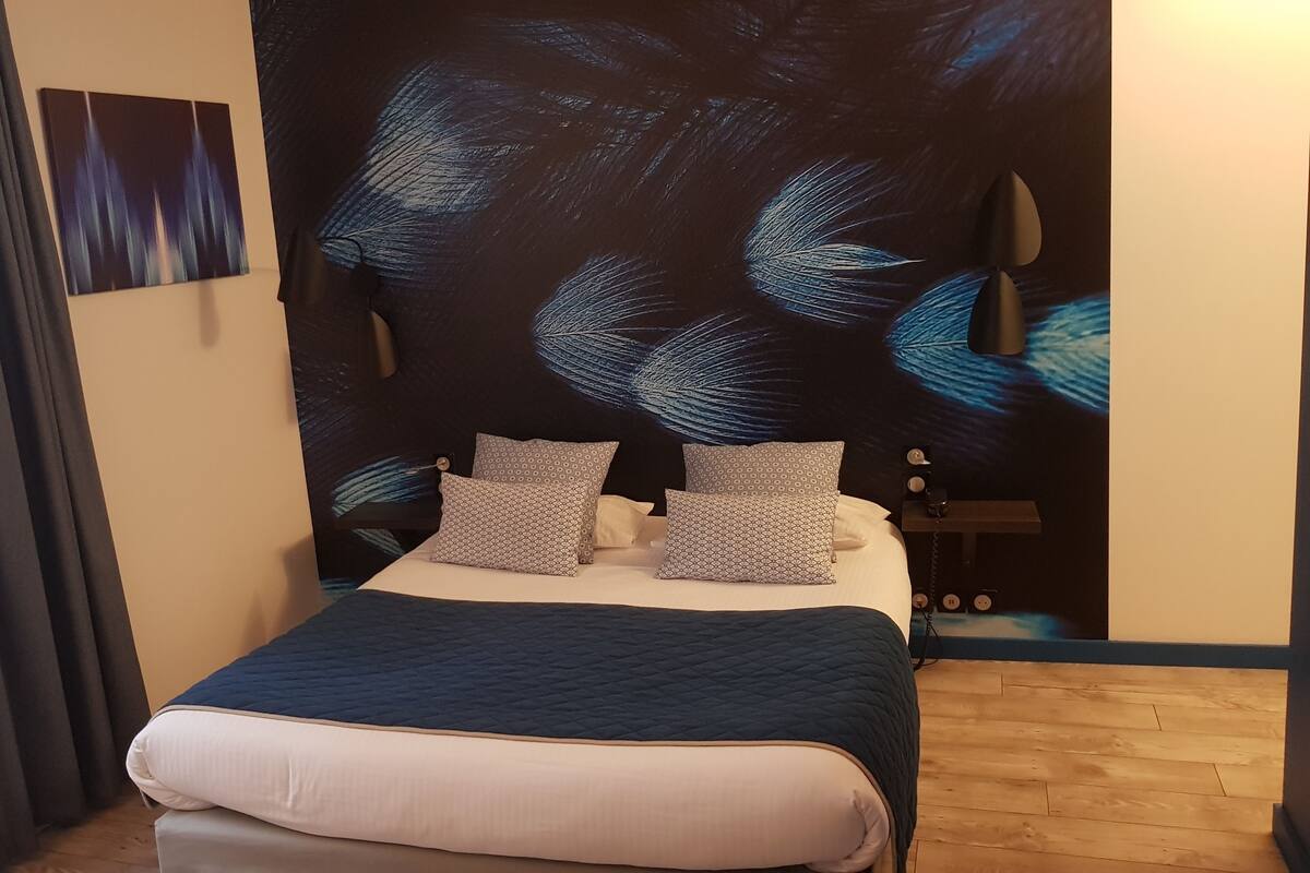 A bed with a dark quilt, patterned pillows, and a wall with a large abstract painting.
