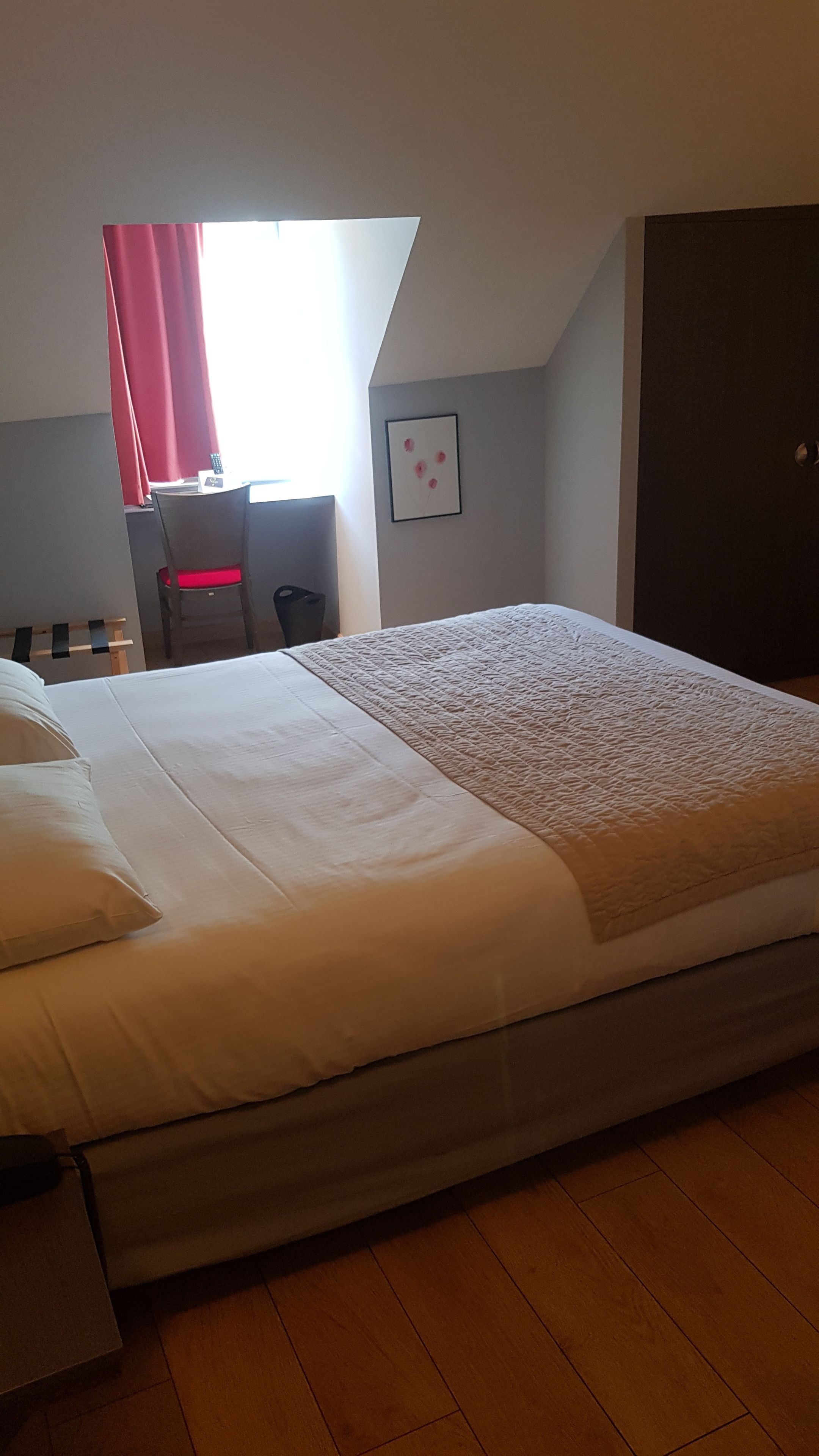 Un dormitorio con una cama, un escritorio y una silla. Hay una ventana con cortinas rojas, un cuadro en la pared y una puerta.
