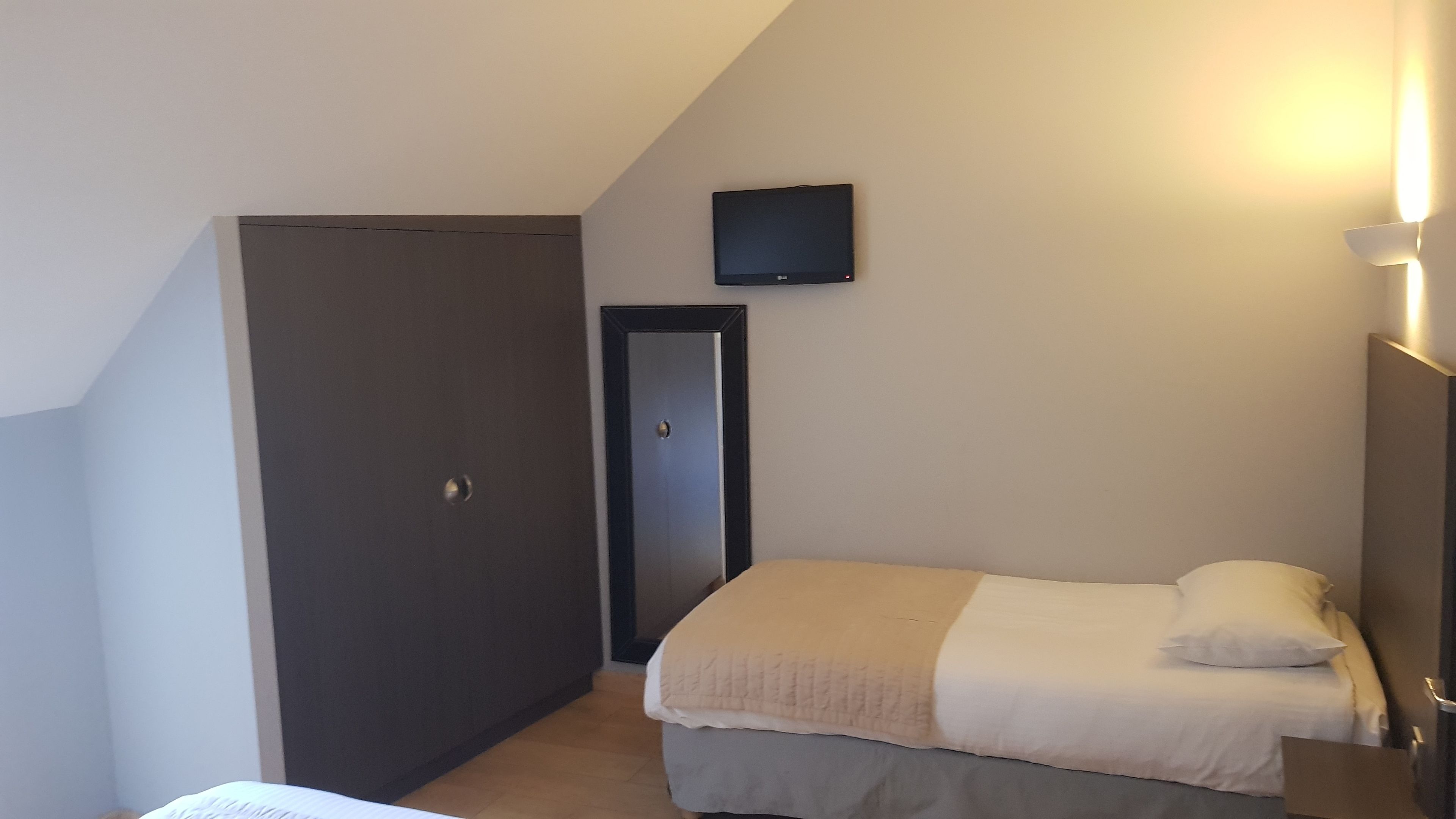 A small, single-bed room with a dark wardrobe, a door, and a wall-mounted TV.