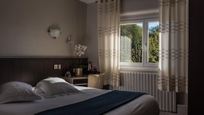 A hotel room with a large bed, a bedside table, a lamp, and a view of trees through the window. - Hôtel CARRER Le Rhien (Ronchamp)
