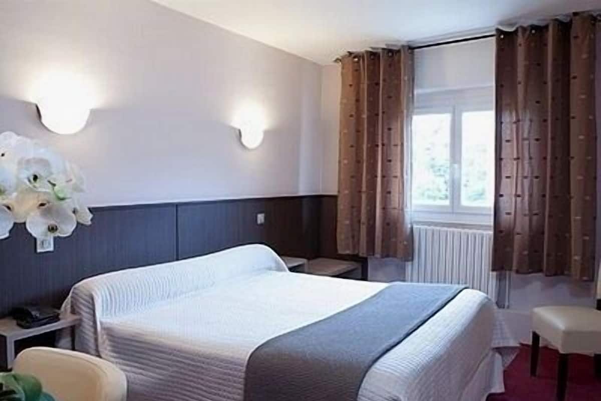 A hotel room with a large bed, a bedside table, a chair, a window with curtains, and a wall-mounted light fixture.