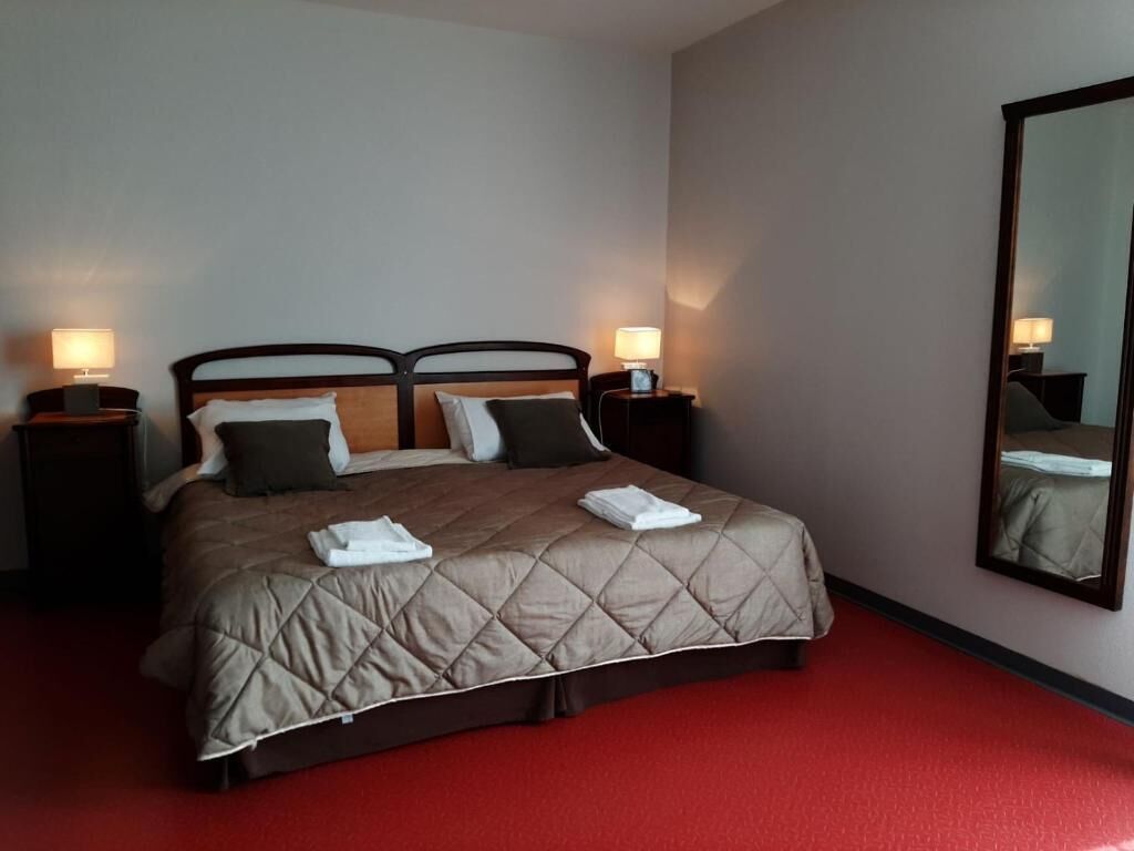 Une chambre d’hôtel avec un lit, deux tables de chevet avec des lampes, un grand miroir et une moquette rouge.