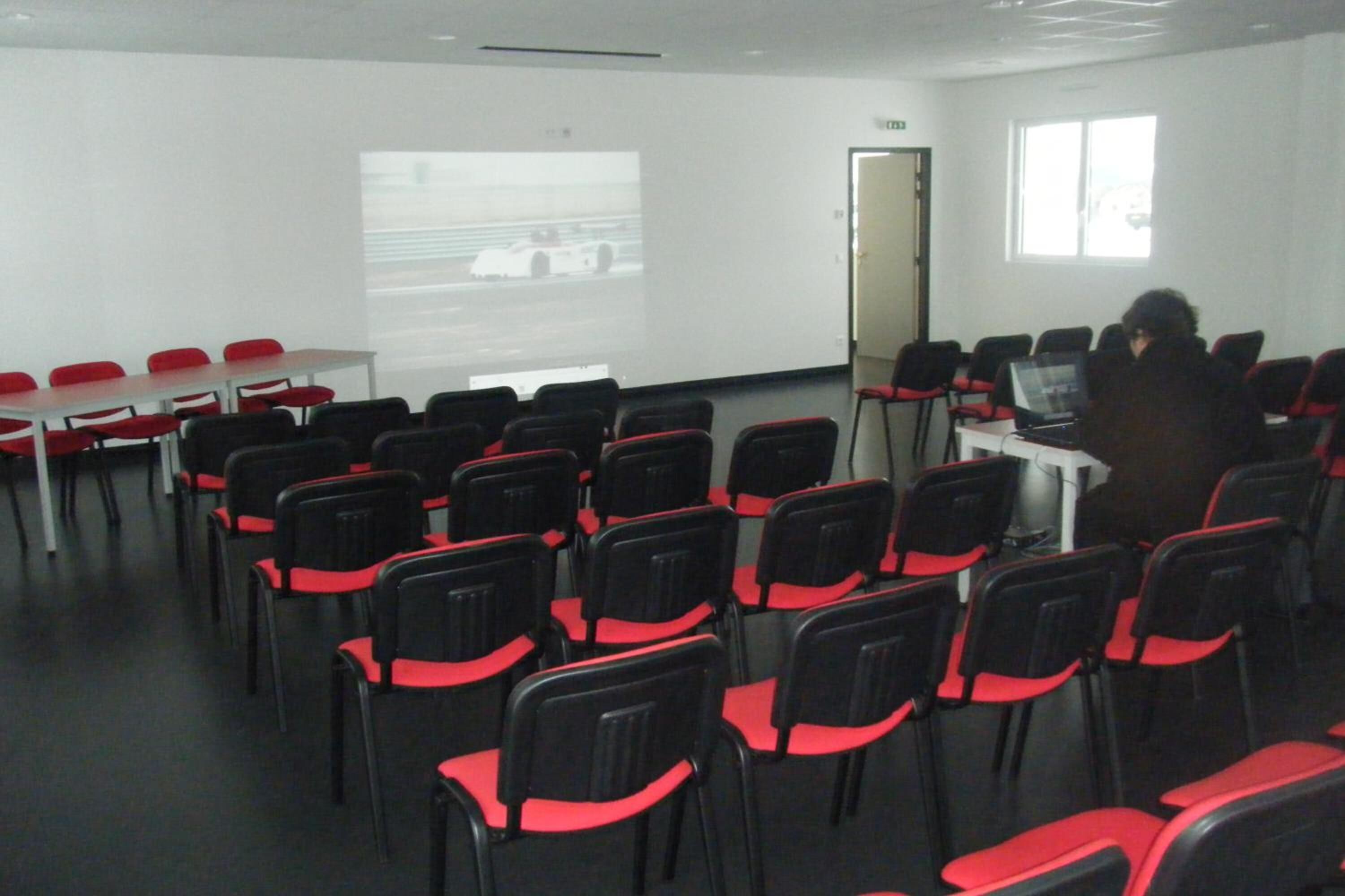 A conference room with rows of black chairs with red accents, a projector screen, and a person at a desk with a laptop.
