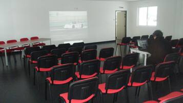 A conference room with rows of black chairs with red accents, a projector screen, and a person at a desk with a laptop.