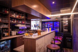 Zona de bar con estantes llenos de botellas de licor, una máquina de café y taburetes de bar.