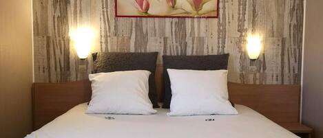 Une chambre d’hôtel avec un lit, deux oreillers blancs, une couvre-lit matelassée de couleur sombre et un tableau représentant des fleurs roses accroché au mur.