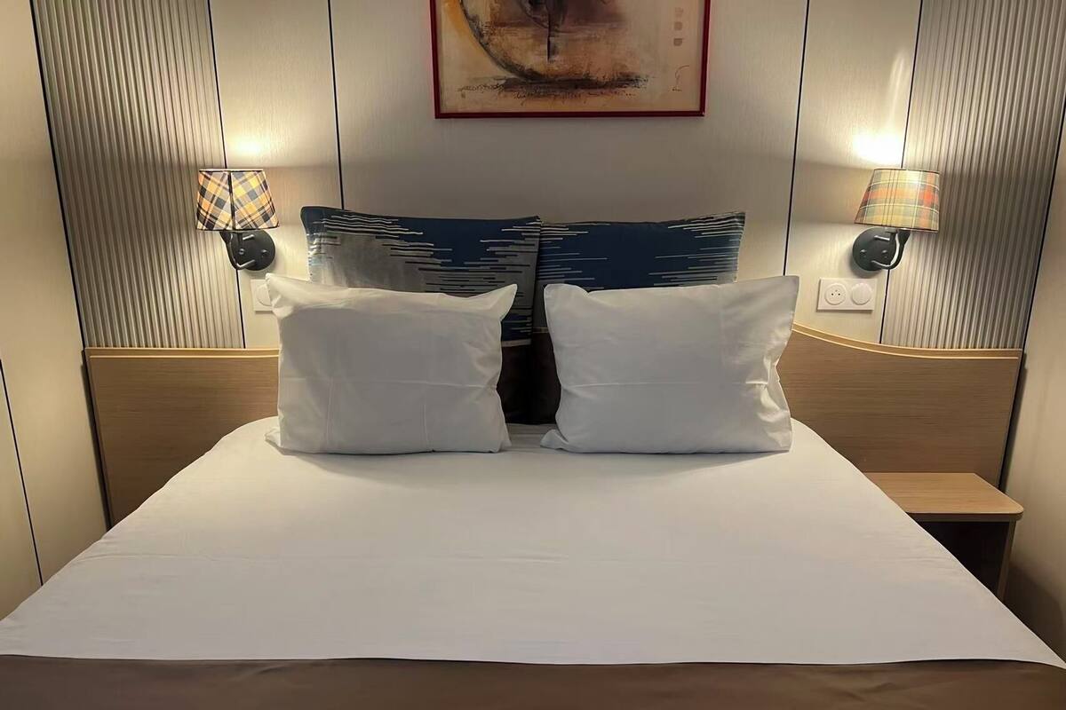 Una habitación de hotel con una cama grande, dos almohadas, una mesita de noche y dos lámparas fijadas a la pared.