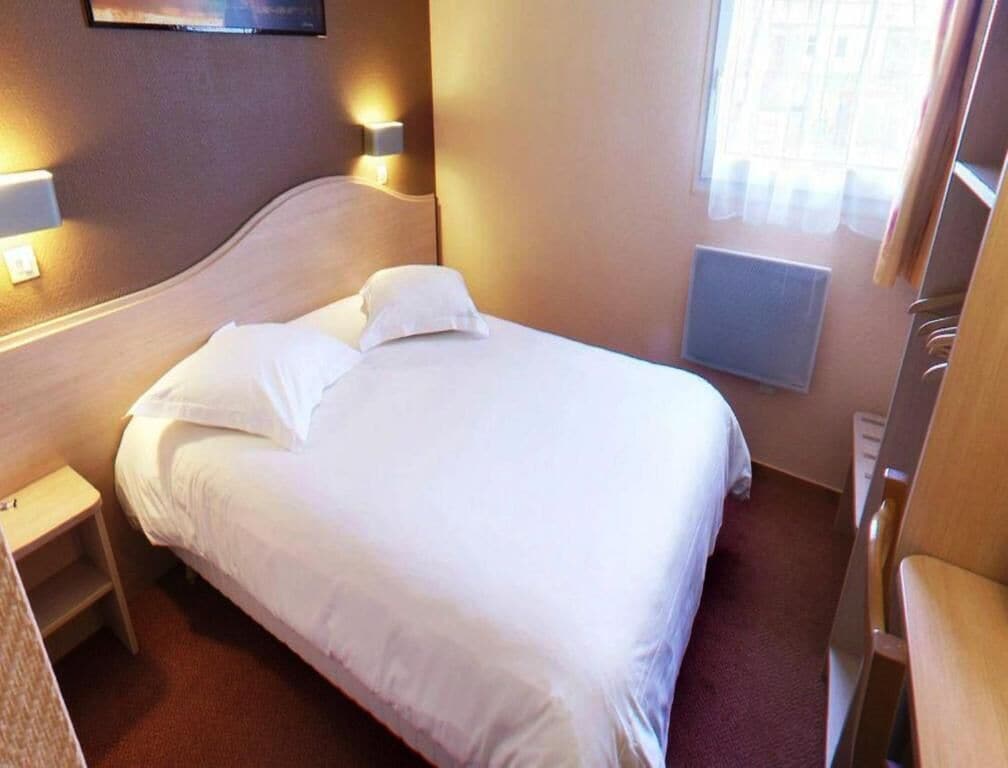 Habitación de hotel con cama, lámparas de noche, mesita de noche, ventana con cortinas y radiador.