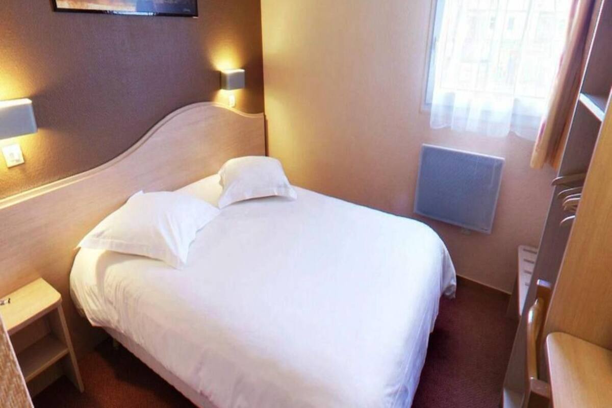 Habitación de hotel con cama, lámparas de noche, mesita de noche, ventana con cortinas y radiador.