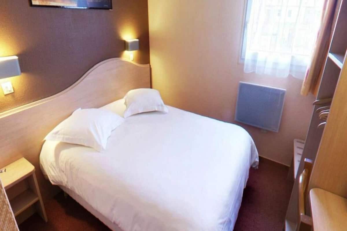 Habitación de hotel con cama, lámparas de noche, mesita de noche, ventana con cortinas y radiador.