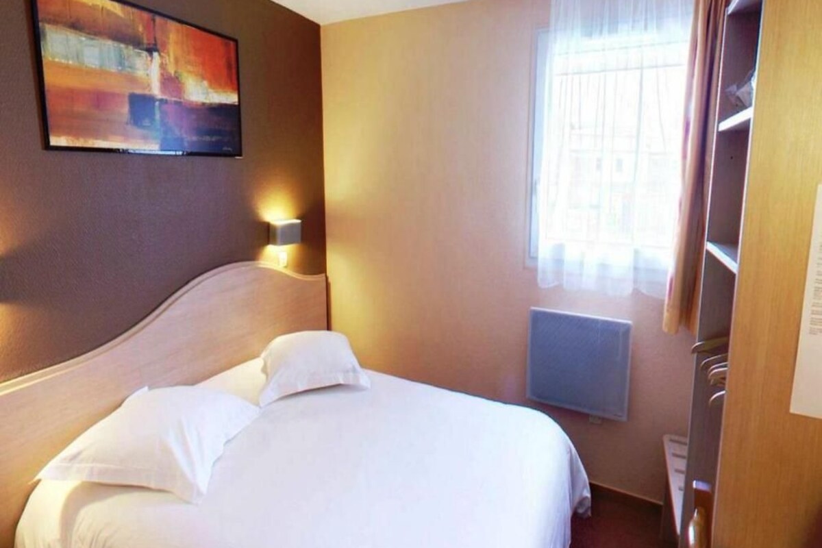A hotel room with a bed, a painting on the wall, a window with curtains, and a radiator.