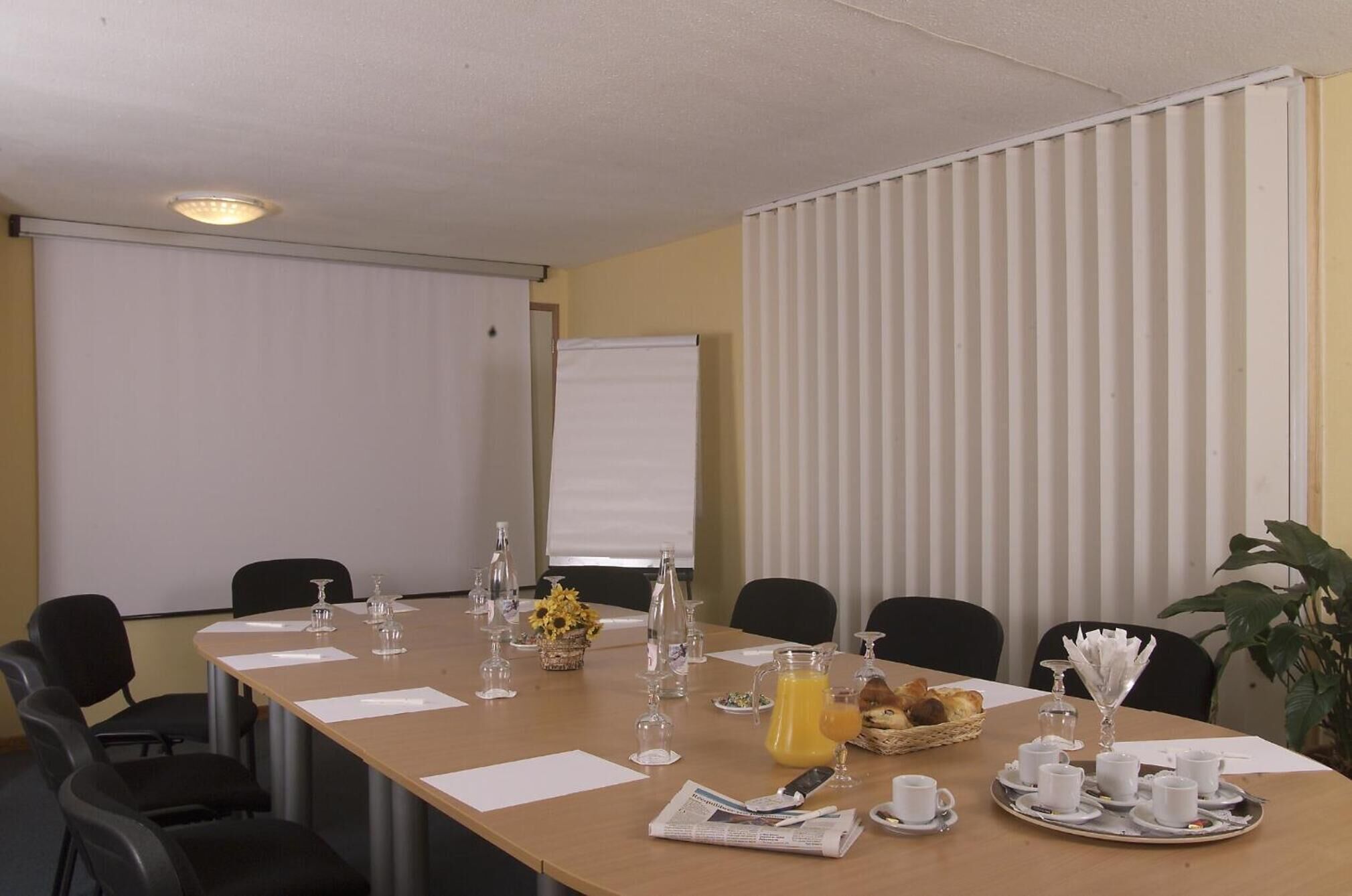 Ein Konferenzraum mit einem langen Tisch, Stühlen, einer Projektionsleinwand und einem Fenster mit Jalousien.